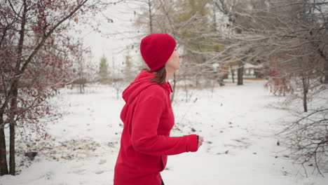 vista lateral de una mujer con gorra roja y sudadera con capucha corriendo al aire libre durante el invierno rodeada de árboles cubiertos de nieve, pinos de hoja perenne y atmósfera brumosa con un edificio borroso en el fondo