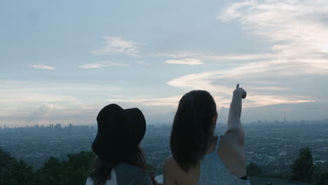 two teenage girls taking a photo and pointing on the sky of a city sunset