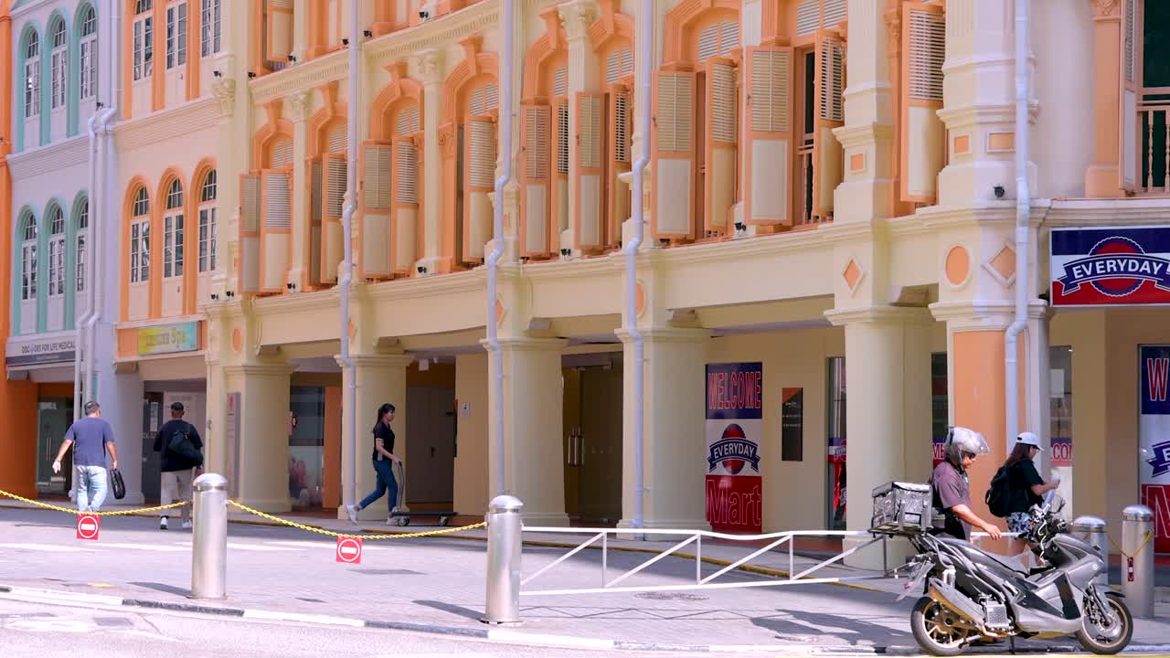 Two motorcyclists ride past vibrant colonial shophouses on a sunny Singapore street, with pedestrians walking by. Bright daylight, static wide shot
