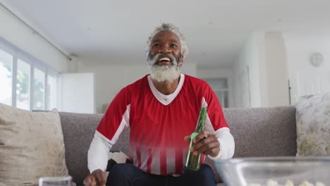 Hombre-Mayor-Afroamericano-En-Casa-Sentado-En-Un-Sofá-En-La-Sala-De-Estar,-Viendo-Deportes-En-La-Televisión,-Dri