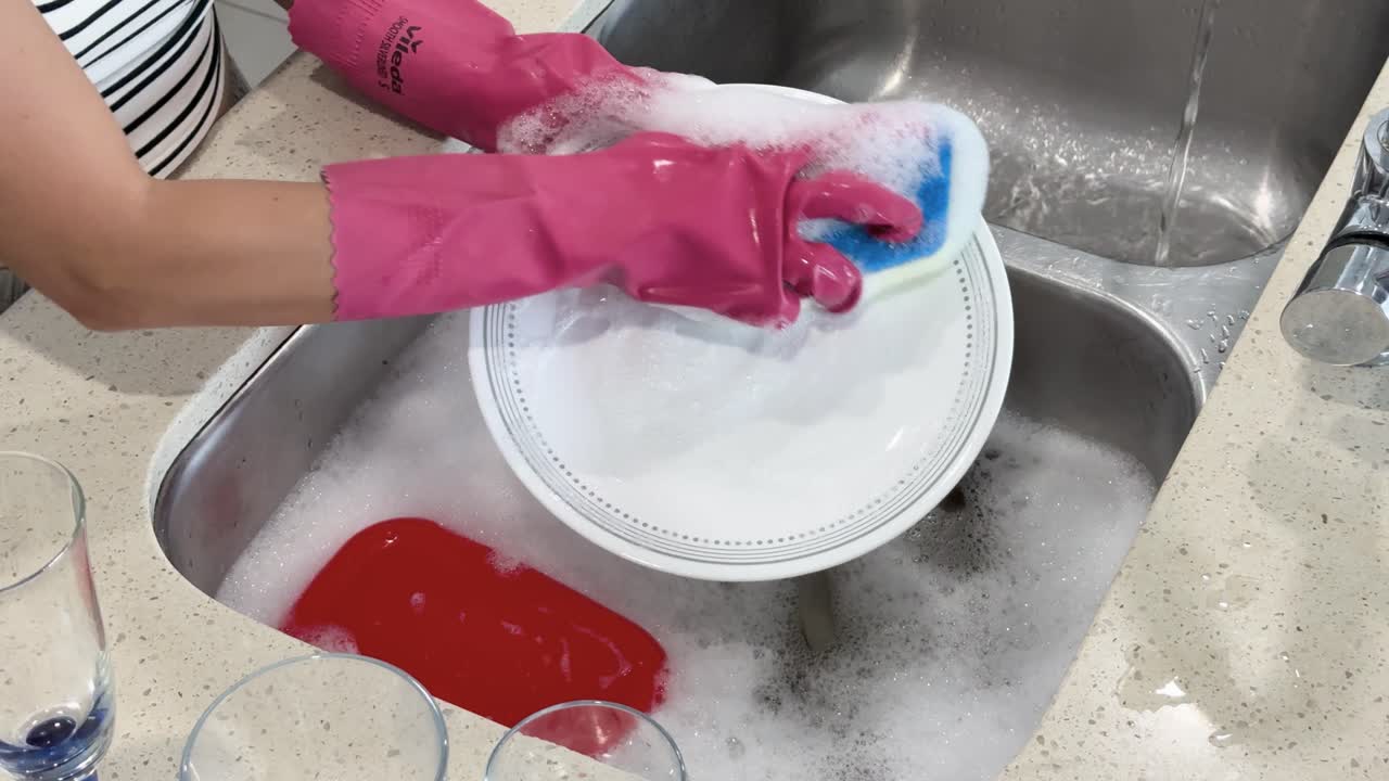 Person wearing pink gloves scrubs and rinses a white plate in a bright kitchen, using soapy water and a sponge, with overhead lighting and steady camera