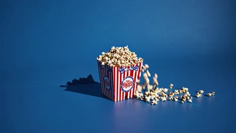 popcorn kernel spilling from overflowing red white striped container, cascading against bright blue backdrop suggesting cinematic excitement and casual snacking