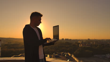 a male stockbroker freelancer stands on a rooftop at sunset with a laptop and types on a keyboard with his fingers looking at the cityscape from a bird's eye view.