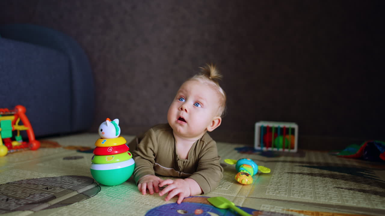 Blond baby boy lies on the floor among the toys. Lovely infant tries to crawl approaching camera.