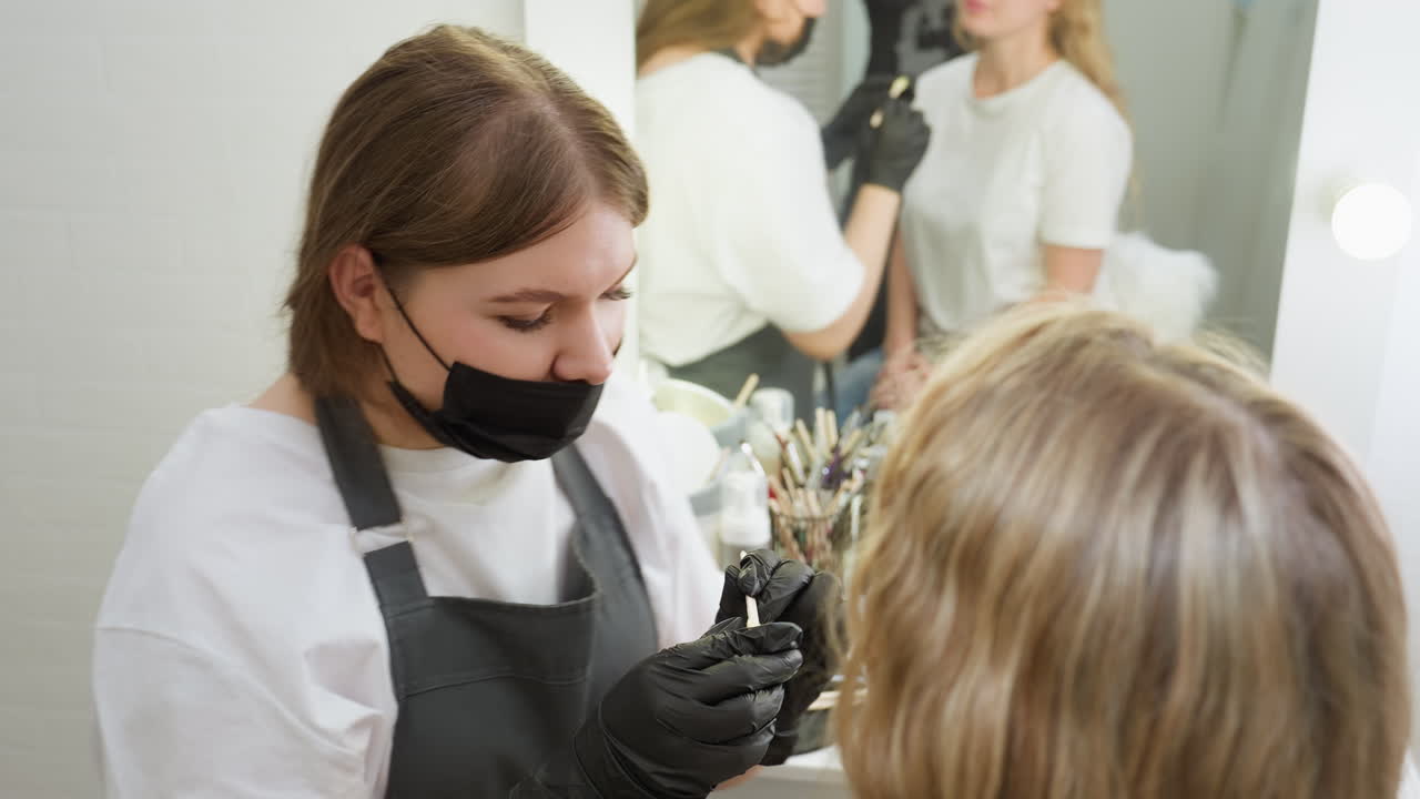 Beautician in mask and gloves focuses while removing wax from client eyebrow in modern salon. Client remains still during facial hair removal as mirror reflects process behind them