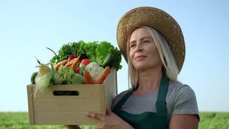 Close-Up-Of-The-Beautiful-Blond-Woman-In-A-Hat-Standing-In-The-Green-Field-And-Holding-A-Box-With-Mature-Vegetables