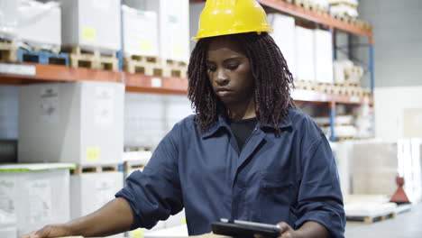 focused african american female employee working in warehouse, checking cardboard box on tablet