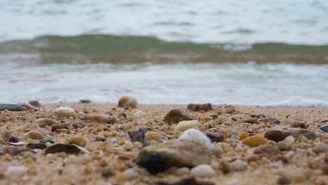 A-low-angle-view-focuses-on-the-foreground,-eroded-seashells-laying-on-the-beach-sand-as-calm-tide-sea-waves-can-be-seen-in-the-background