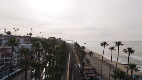 Vista-Aérea-De-La-Ciudad-Costera-De-California-Con-Surfistas-Cruzando-El-Puente-Sobre-Las-Vías-Del-Tren.