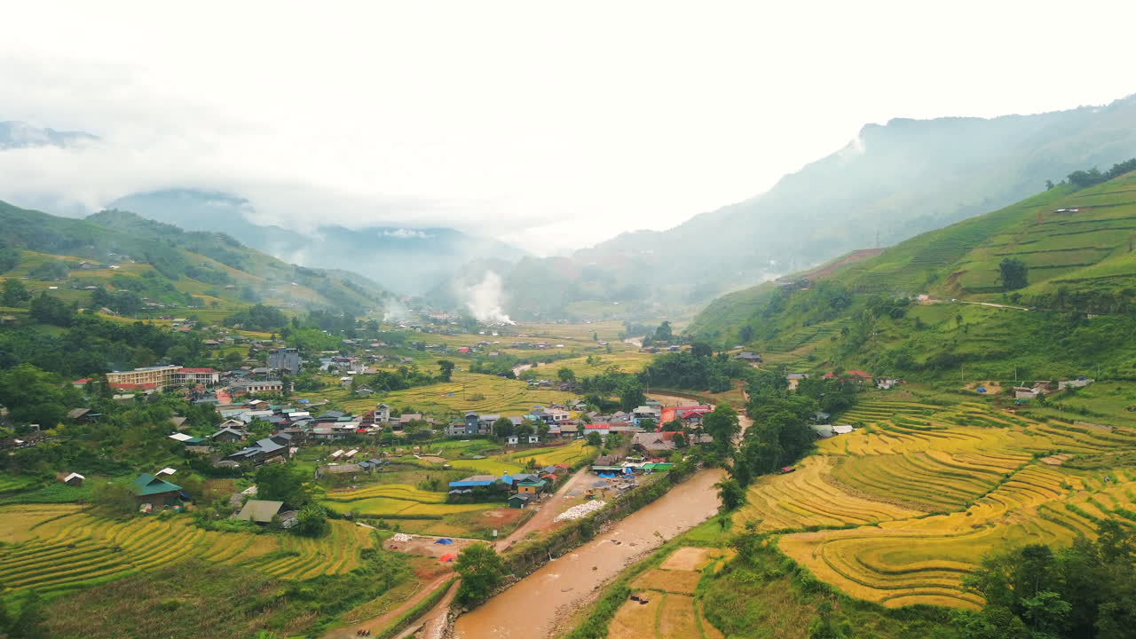 Tangkapan drone di atas lembah yang indah di Sa Pa, Vietnam, menampilkan sawah terasering keemasan, sungai berwarna cokelat, dan rumah-rumah tradisional di bawah langit gunung yang berkabut.