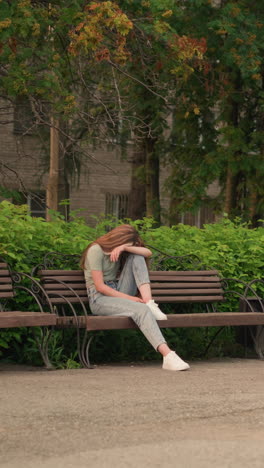 joven sentada en un banco de madera en un parque, apoyando su cabeza en su brazo en una postura reflexiva y sombría, rodeada de exuberante vegetación, árboles y una figura de fondo que se mueve cerca