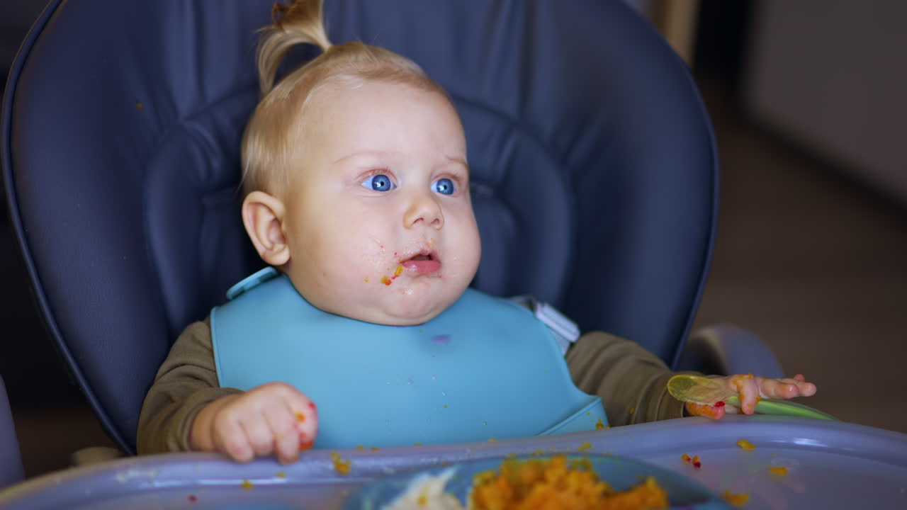 Cute blond baby with a bib sits in high chair. Adorable infant holds a spoon in his hand. Close up.