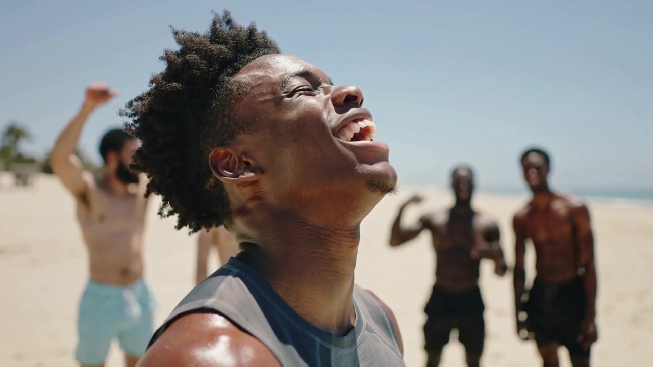 Wiping sweat from sun, Black man in sleeveless top exhaling and laughing on beach, friends cheering