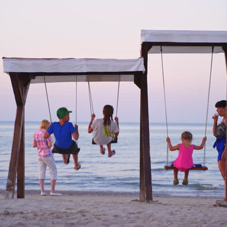 children having fun on wooden swings on the sea background. playground on sandy beach and children are swinging in the evening, looking at sea water.
