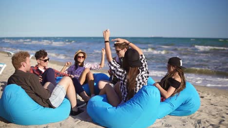 Young-man-playing-guitar-among-group-of-friends-sitting-on-easychairs-on-the-beach-and-singing-on-a-summer-evening.-Slowmotion-shot