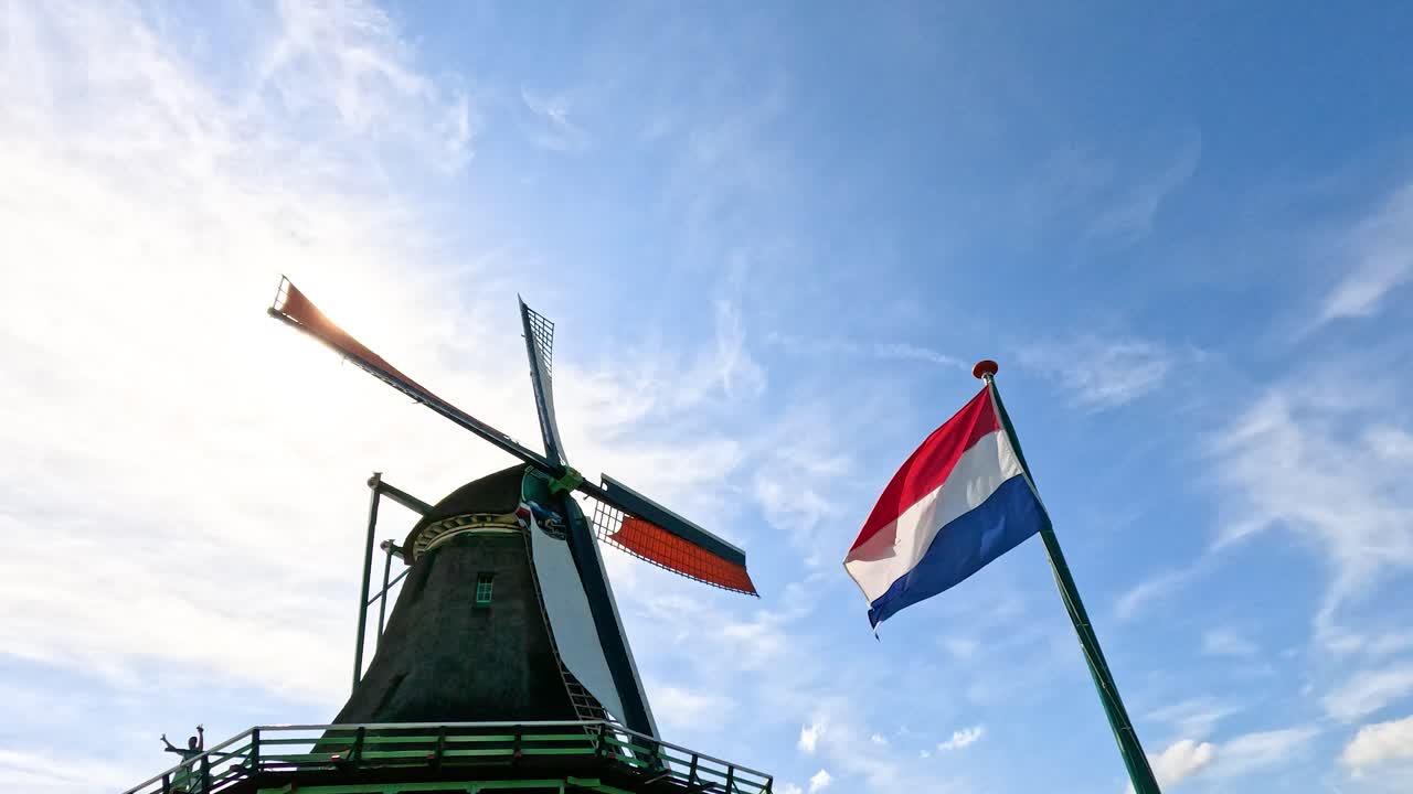 Historic windmill blades rotate beside Dutch flag, bright daylight, clear blue sky, low angle