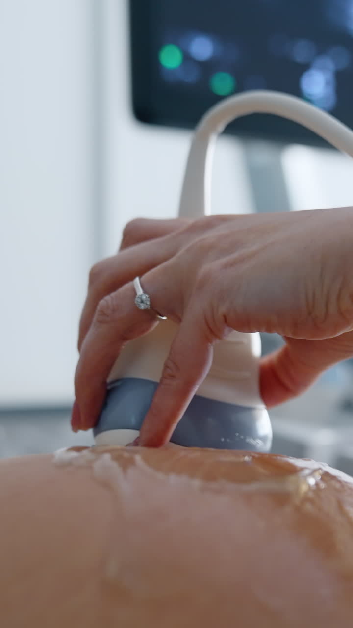 Female hand sliding the ultrasonic transducer by the round pregnant belly. Gel is spread by the female abdomen. Close up. Vertical video