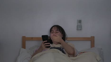 woman sits in bed holding phone, tired, face illuminated by phone screen