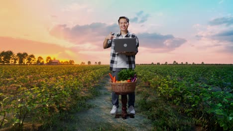cuerpo lleno de granjero masculino asiático con canasta de verduras sonriendo mientras usa y apunta a la computadora portátil en el campo