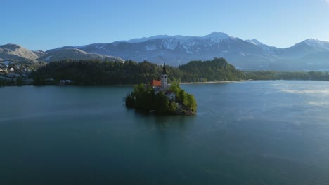 Picturesque-church-on-an-island-in-the-middle-of-a-serene-lake,-surrounded-by-mountains-and-trees-under-a-clear-sky