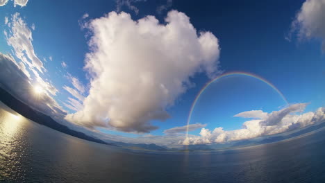 stunning wide-angle view of a rainbow over a lake with clouds and sun