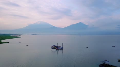 Aerial-view-of-dredger-boat-with-excavator-on-the-lake-to-clean-up-trash-and-scrape-mud-with-view-of-mountain-on-the-background---Rawa-Pening-Lake,-Indonesia