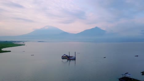 aerial view of dredger boat with excavator on the lake to clean up trash and scrape mud with view of mountain on the background - rawa pening lake, indonesia