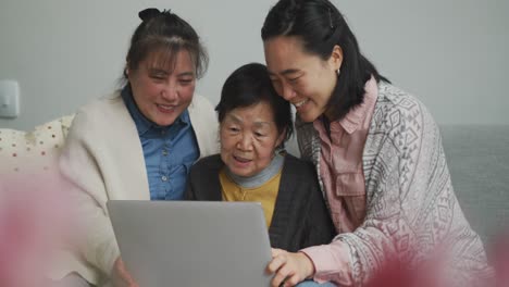 Happy-senior-asian-mother,-adult-daughter-and-granddaughter-in-living-room-using-laptop,-embracing