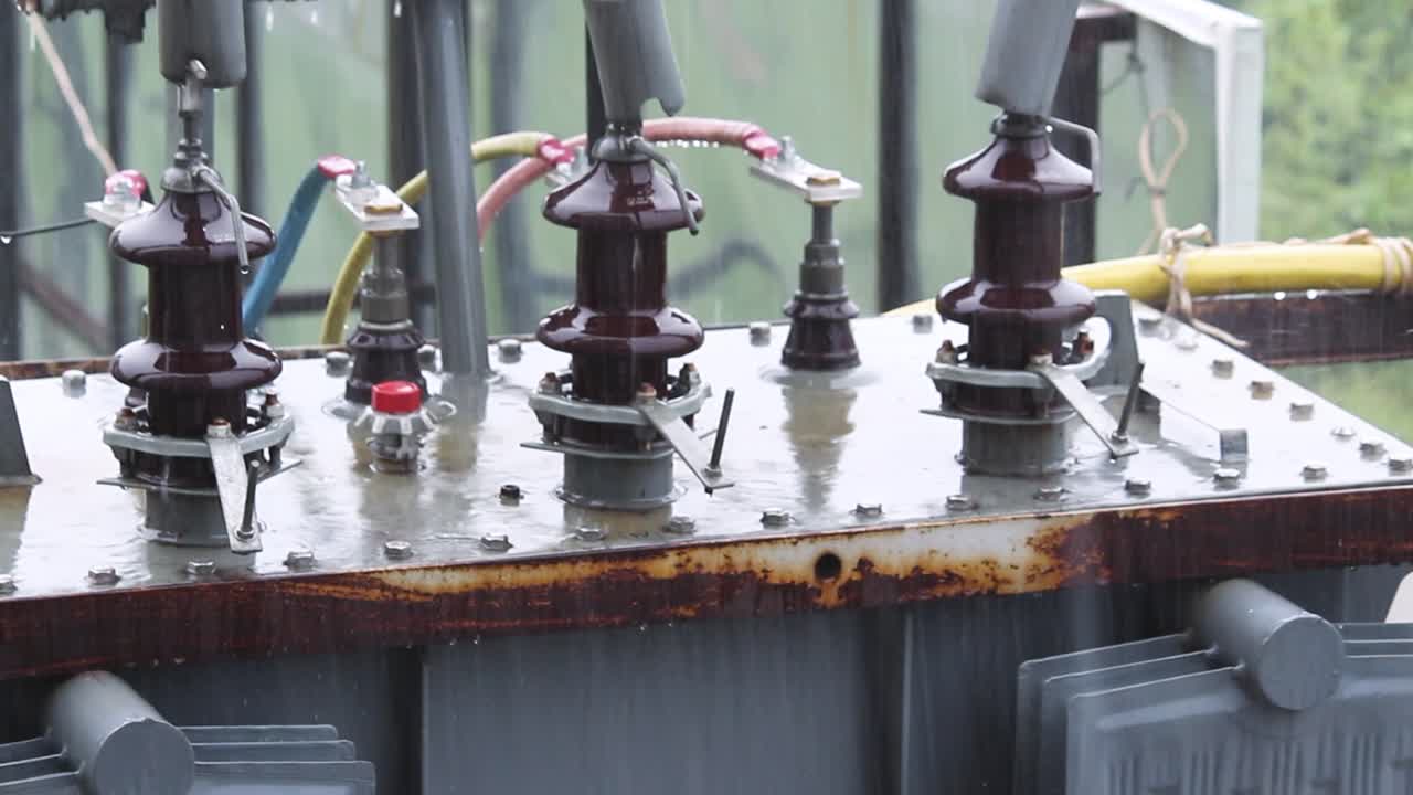 Electrical transformer closeup under rain, high voltage insulators, wires and rusted surface during monsoon. Ideal for energy, industry or weather-related footage