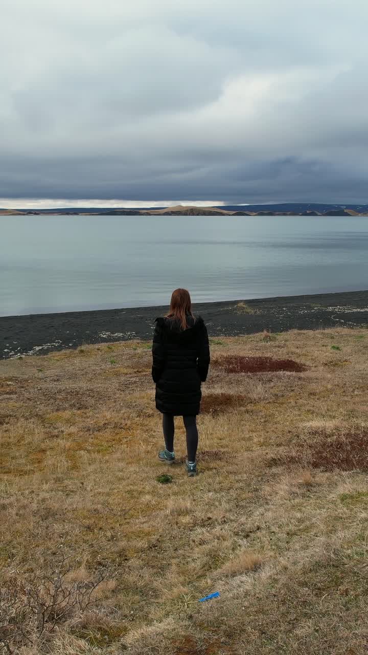 Mujer caminando por un campo hacia un lago con una playa de arena negra en un día nublado en Islandia