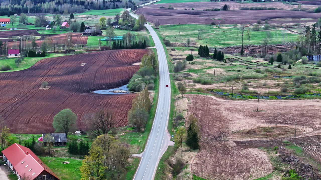 Static aerial view shows a two-lane asphalt road with cars driving through a rural Latvian village surrounded by plowed agriculture fields and trees
