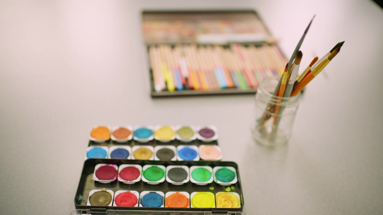 Close up of paintbrushes in a glass jar with watercolor palette and colored pencils blurred in the background