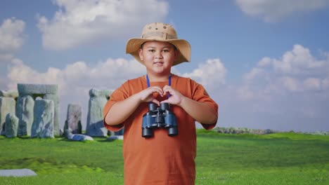 asian boy with a hat and binoculars making heart shape gesture while traveling in stonehenge. boy researcher examines something, travel tourism adventure concept
