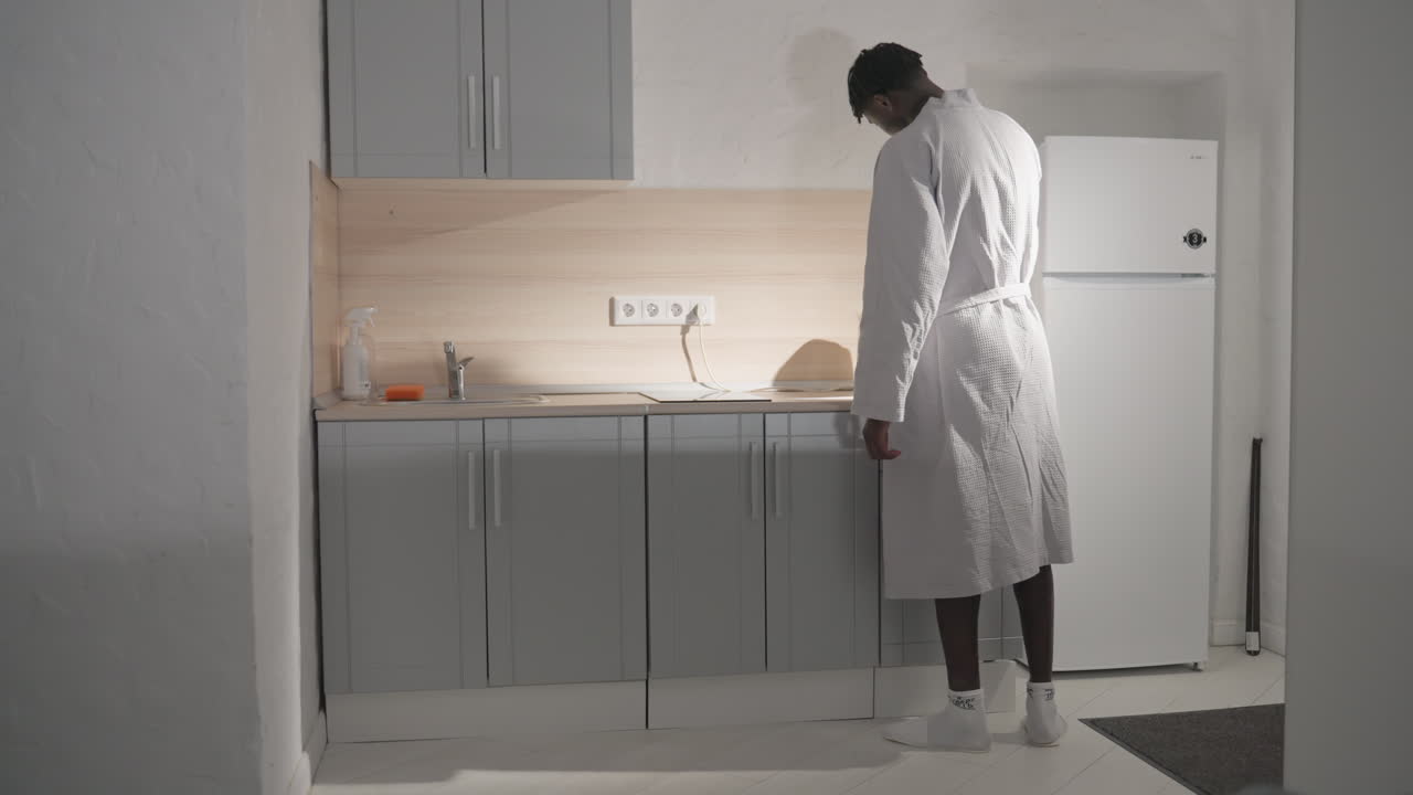 Man in white bathrobe preparing coffee in modern kitchen. He stands by counter, holding electric kettle while the minimalist kitchen is bathed in soft morning light. Neat, contemporary kitchen space