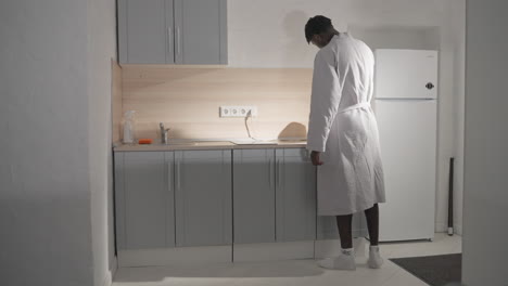 man in white bathrobe preparing coffee in modern kitchen. he stands by counter, holding electric kettle while the minimalist kitchen is bathed in soft morning light. neat, contemporary kitchen space
