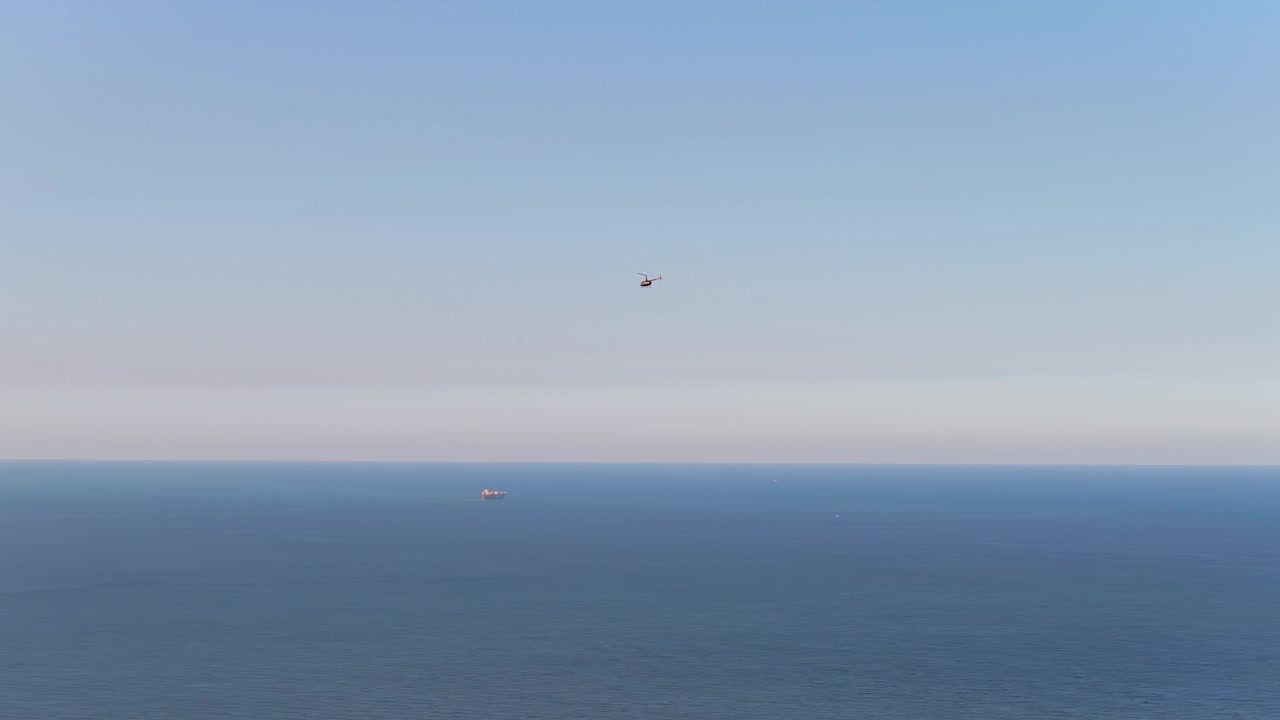 En helikopter svæver over det vidtstrakte ocean, mens skibe prikker horisonten. Fanger en følelse af isolation og uendelighed. Velegnet til maritime eller luftfartstemaer.