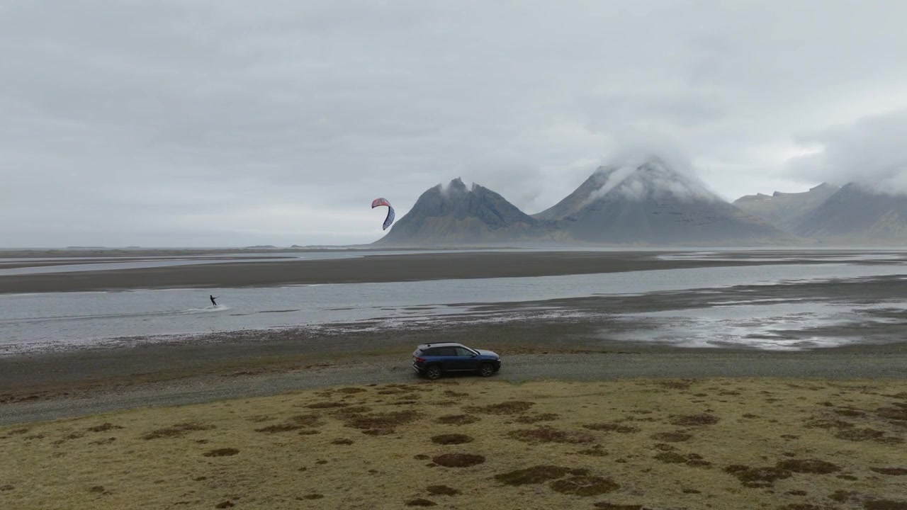 SUV drives across the black sand at Stokksnes near Hofn, Hornafjordur, Iceland, while a kitesurfer rides beneath the cloud covered Vestrahorn massif, drone tracking shot