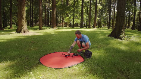 Man-packs-up-small-drone-after-flight-in-park,-sunny-day,-wide-shot