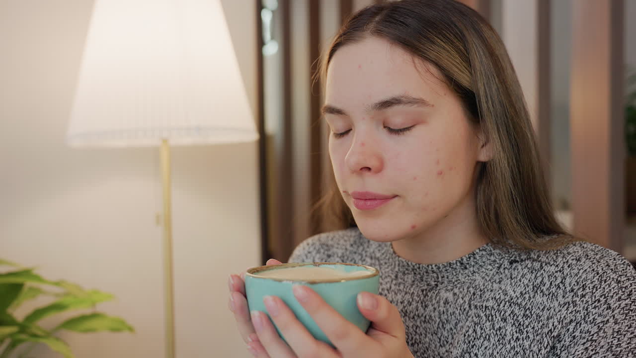 Young lady gently smelling steam from hot tea held in both hands with satisfaction on face seated indoors in cozy setting soft light from lamp nearby potted plant in background serene peaceful