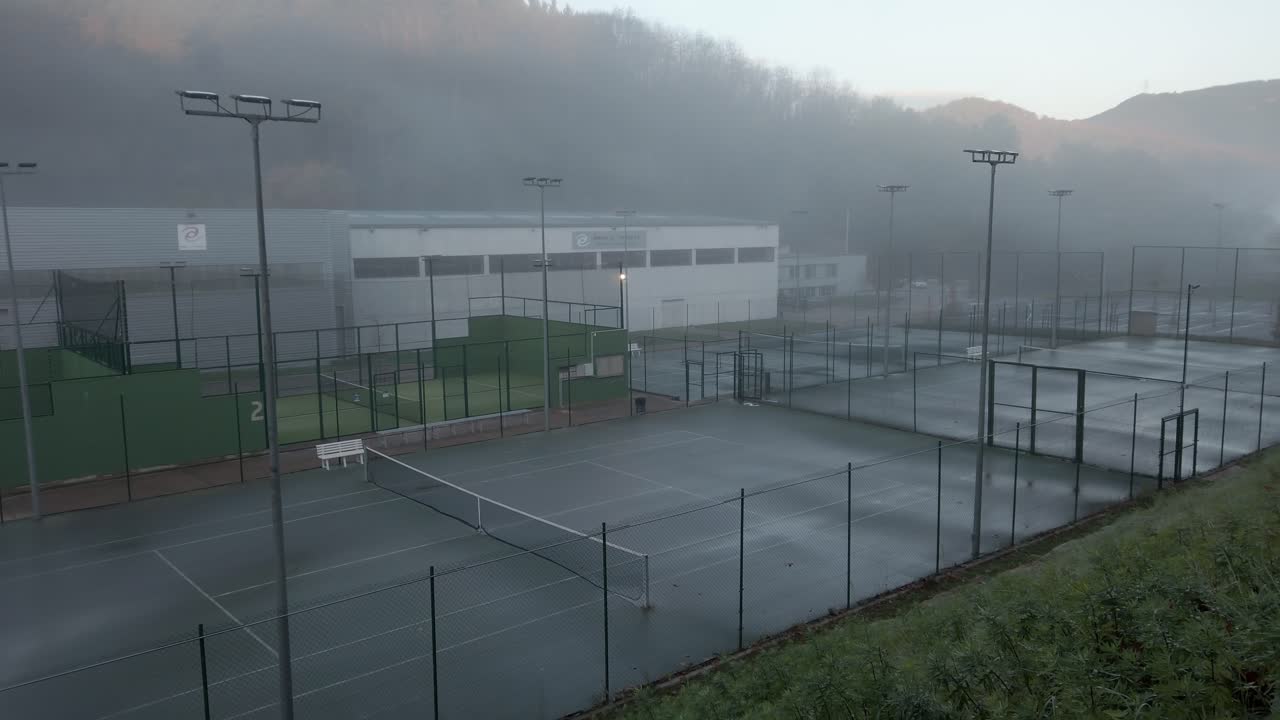 Sunlight illuminates fog rolling over several tennis courts and paddle courts next to a large building in the mountains of catalonia, spain