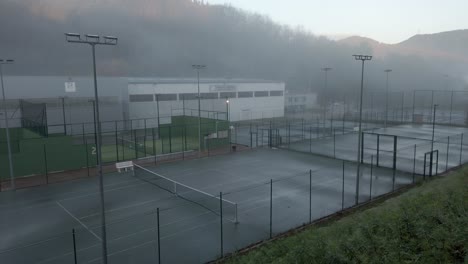 sunlight illuminates fog rolling over several tennis courts and paddle courts next to a large building in the mountains of catalonia, spain