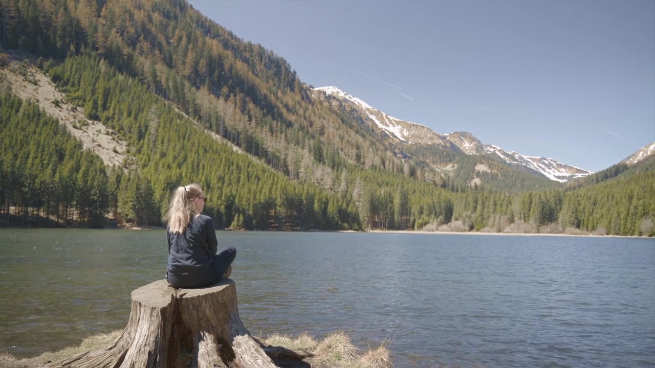 Vista posteriore di una ragazza caucasica persa in contemplazione in una giornata di sole di fronte a un paesaggio montano scenografico, calmo e tranquillo, acqua di fiume che scorre e una leggera brezza.