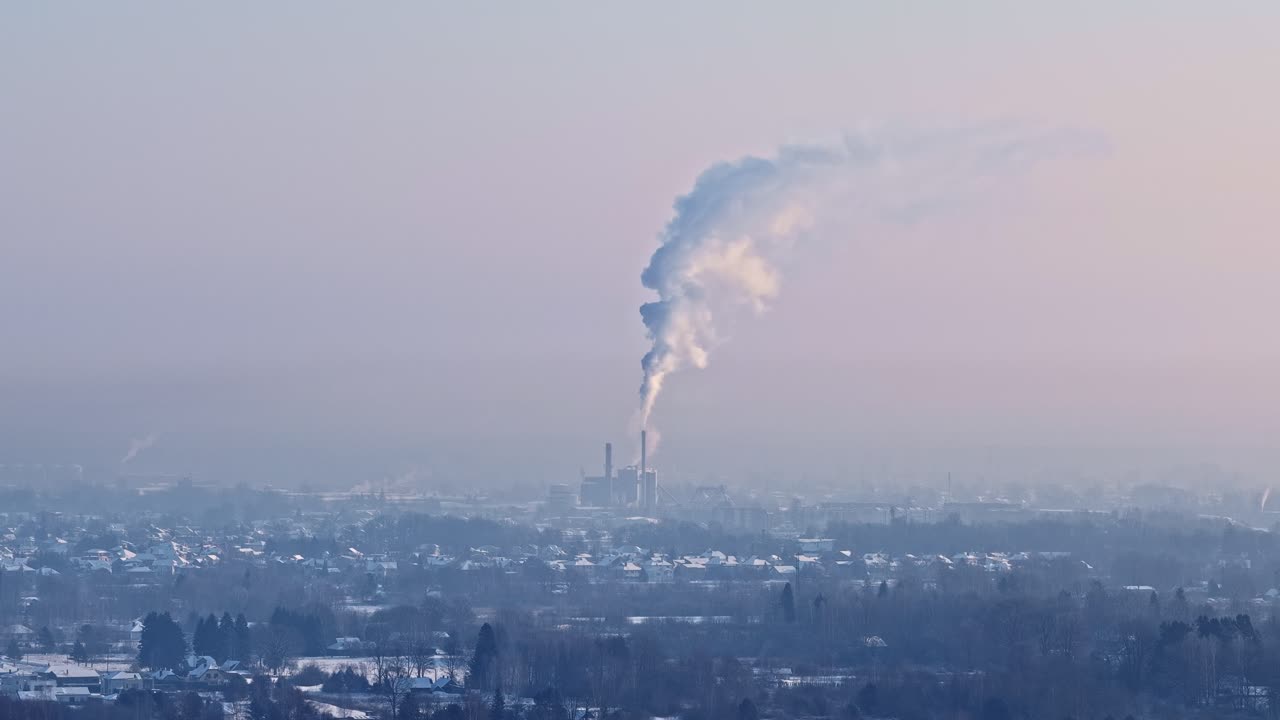 Alba: ciminiere distanti di un impianto industriale rilasciano un grande pennacchio sopra lo skyline nebbioso della città, mostrando l'inquinamento atmosferico urbano e le emissioni contro un cielo color pastello