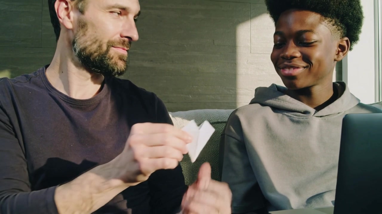 Opening paper, adult male guiding teen's fingers on couch, teaching game with white fortune teller