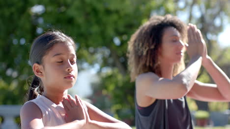 Feliz-Madre-Birracial-E-Hija-Practicando-Meditación-De-Yoga-Sentadas-En-Un-Jardín-Soleado,-Cámara-Lenta