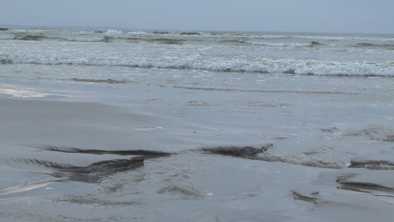 Static view of sea shoreline with white wet sand and gray blue sky. Relaxing slow motion of foamy waves roll in to the beach, Estonia coast. Low angle view waves are reaching the shore on overcast day