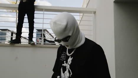 guy in a white ski mask, chain and sunglasses acting tough with guy in the background standing on rails