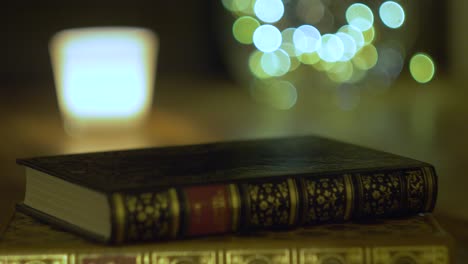 Old-Book-Lying-On-A-Wooden-Table-With-A-Bright-Bokeh-Balls-And-A-Candle-In-The-Background-in-interior-of-house-with-vintage-and-festive-mood