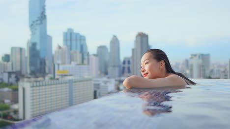 a pretty young woman leans on the edge of a rooftop pool looking out over a modern city skyline