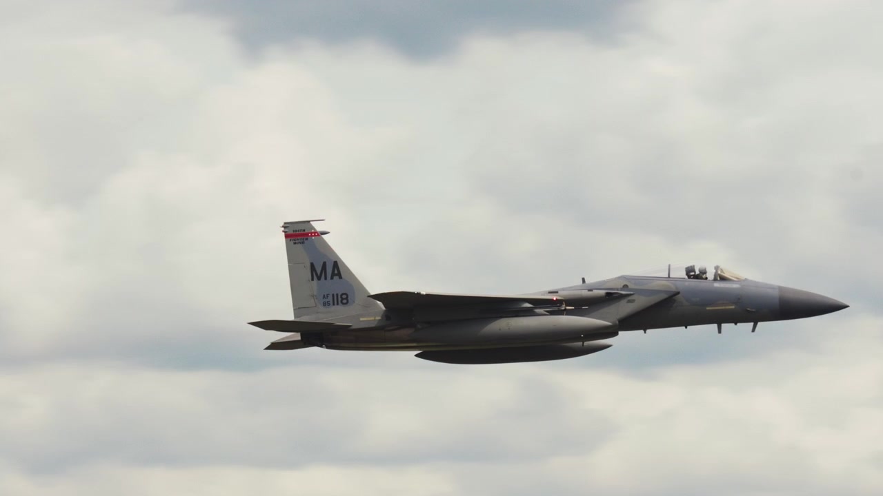 An F-15 military fighter jet banks and climbs through an overcast sky.